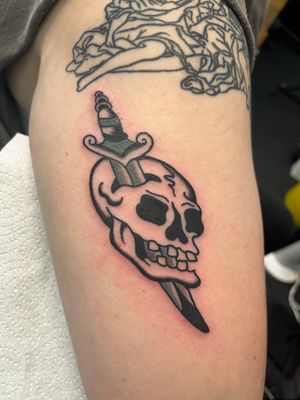 Get inked with a classic traditional tattoo featuring a fierce skull and menacing dagger, expertly done by Claudia Vicente.