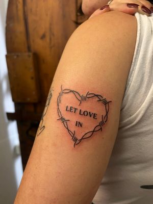 Get inked with a small lettering heart tattoo featuring the quote 'Let Love In' by Claudia Vicente. An illustrative design that spreads positivity.