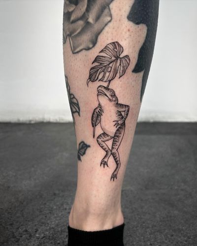 Experience the intricate beauty of dotwork in this unique tattoo featuring a frog and monstera leaves, created by the talented artist Paula.