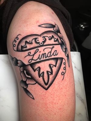 Small lettering and traditional style black and gray tattoo featuring a heart and ribbon design by Angel Face.