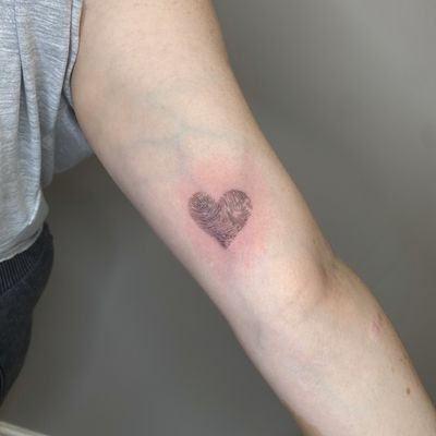Express your unique love story with a delicate fine line tattoo of a heart intertwined with a fingerprint by Tas Kal.