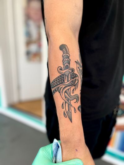 Explore the fierce combination of a snake and dagger in this classic traditional style tattoo by River Tatts.