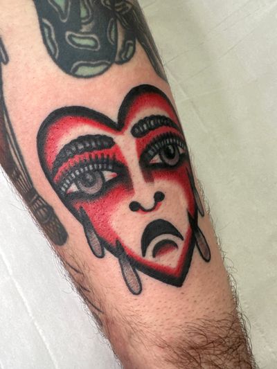This traditional tattoo features a sad face within a heart motif, expertly executed by artist Jakob Isaac. A timeless design with emotional depth.