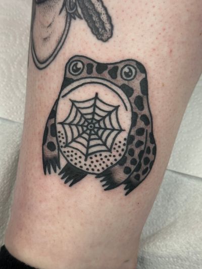 Experience the mystical allure of nature with this traditional tattoo featuring a frog and spiderweb, expertly done by Jakob Isaac.
