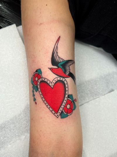 Get a classic traditional tattoo featuring a beautiful bird, flower, and heart design by the talented artist Ryan Goodrum.