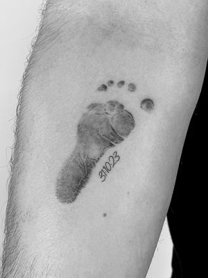 Small lettering tattoo of a baby's footprint, done in black and gray by Saka Tattoo.