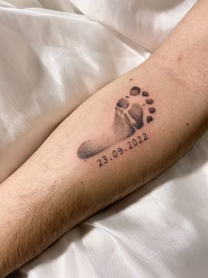 A beautifully detailed black and gray tattoo featuring a baby footprint and a date, done in small lettering in an illustrative style by the talented artist Gabriela.