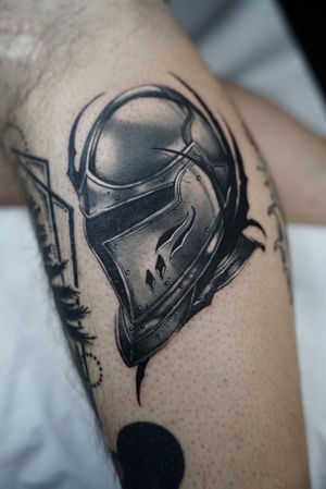 Experience the intricate detail and fierce symbolism of a black and gray knight helmet tattoo, expertly crafted by renowned artist Dominga Longo.