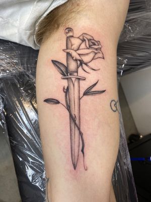 Illustrative black and gray tattoo of a flower intertwined with a dagger, expertly done by artist Charlie Macarthur.