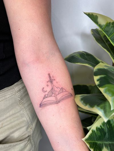 Immerse yourself in a world of imagination with this fine line, illustrative tattoo by Emma InkBaby featuring mountains, a sword, and a mystical book.