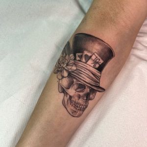 Illustrative black and gray tattoo featuring a skull wearing a top hat surrounded by flowers and playing cards, designed by Cristina.