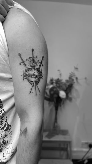 Experience the powerful symbolism of a black and gray illustrative tattoo featuring a sacred heart entwined with a sword, expertly crafted by Rollo.