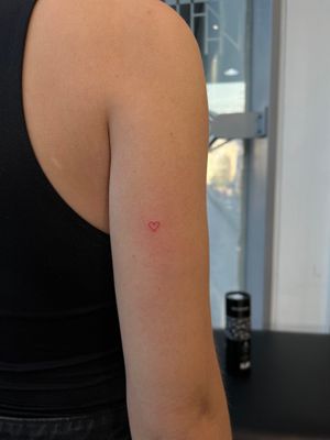 Express your love with this delicate fine line heart tattoo in vibrant red ink by the talented artist Faith Llewellyn.