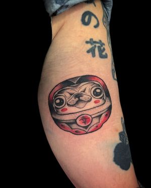 Get a playful and adorable Japanese style tattoo of a pug transforming into a Daruma doll, expertly done by artist Nat.