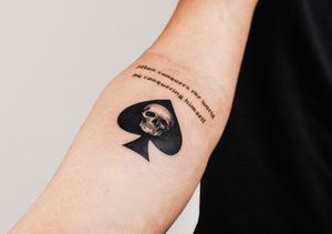 Small lettering and illustrative style tattoo featuring a skull and spades motif by Gabriele Edu.