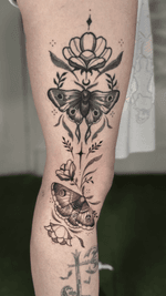 A blackwork tattoo extending vertically on the leg, featuring two detailed moths surrounded by various blooming flowers, stylized leaves, dotwork accents, and star-like abstract elements, creating a cohesive and mystical design.