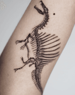 A black ink tattoo of a spinosaurus skeleton, running down the arm. The tattoo emphasizes the skeletal structure with detailed lines and shading. The style seems anatomical with a touch of artistry.