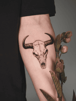 A black and grey tattoo depicting the skull of a bull. The style is detailed with fine shading and lines, emphasizing the cracks and textures of the skull.