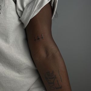 Minimalist blackwork tattoo by 'Scary' Tate Hazen featuring small lettering of a number on dark skin.