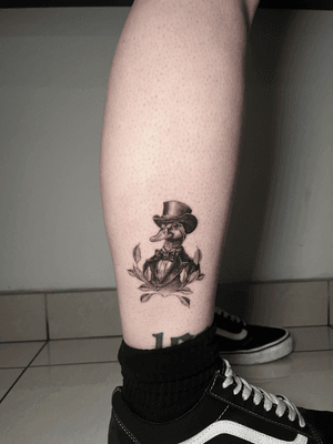 A detailed black and gray tattoo featuring a gentleman wearing a top hat, alongside a charming duck. Created with micro realism and realism techniques by artist Lauren.