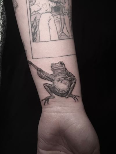 Capture the harmony of nature with this black and gray illustrative tattoo by Lauren. A whimsical frog playing a banjo will bring a touch of music and magic to your skin.