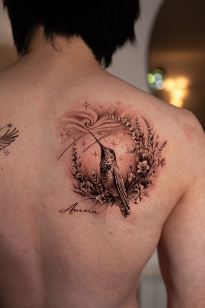 Experience the beauty of micro-realism with this stunning black and gray tattoo by Ion Caraman, showcasing a hummingbird drinking nectar from a flower surrounded by a crown, vine, and stars.