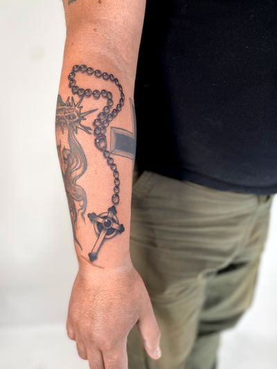 A black and gray illustrative tattoo of a cross intertwined with a rosary, created by tattoo artist Alfonso Barberio.