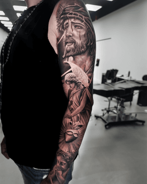 A beautiful black and gray realism tattoo featuring Jesus, an angel, a sacred heart, a praying child, all expertly done by Ember.