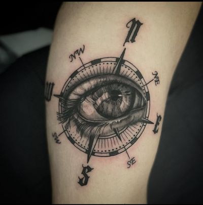 This black and gray tattoo by Cara Hayward features a realistic compass, abstract eye, and small lettering, symbolizing travel and exploration.