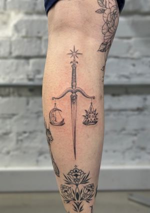 Elegant black and gray tattoo by Ellie Shearer featuring a sun, moon, star, sword, and scale symbolizing cosmic harmony.