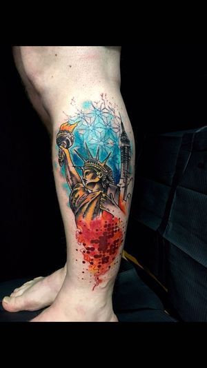 A stunning lower leg tattoo by Sandro Secchin featuring a torch and architectural elements in a vibrant geometric watercolor style.