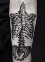 A detailed blackwork tattoo depicting a human rib cage, spine, and pelvic bones, rendered with textured lines, extends along the forearm.