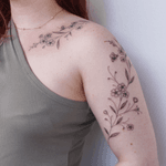 A delicate floral design tattoo that wraps along the shoulder and arm. It features forget-me-not flowers. The style of the tattoo is minimalistic and elegant in a feminine fineline style