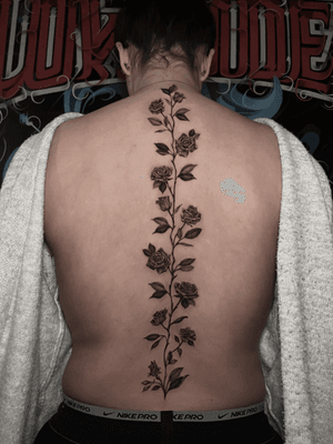 Get a stunning black and gray illustrative tattoo of a rose vine along your spine by the talented artist Lauren.