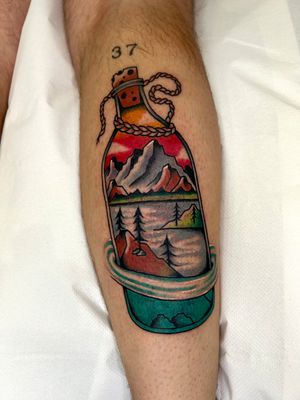 A neo-traditional tattoo depicting a serene landscape with mountains, a lake, forest, and a bottle. Created by Barney Coles.