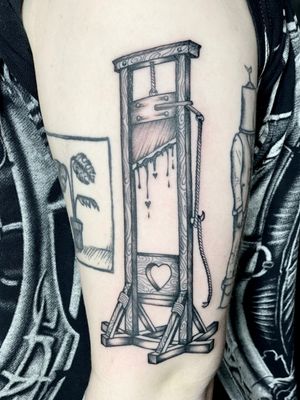Black and gray tattoo of a guillotine by artist Amandine Canata, featuring detailed illustrative style.