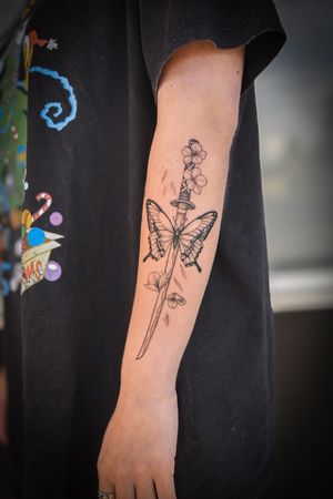 Admire the delicate details of this black and gray fine line tattoo featuring a butterfly, flower, and sword, by talented artist Steffan Eagle.