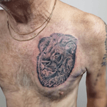 A black and white tattoo of a lion's head in a realistic style. The lion's expression is serious. The details and shading suggest the use of stippling or fine lines to give texture.