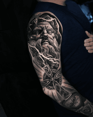 A black and gray tattoo by Ember, featuring a rose, compass, cross, Odin, Zeus, lightning, and knot in a realistic style.