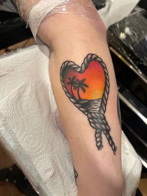 Get lost in the beauty of this traditional tattoo featuring a serene sunset, beach scene, palm tree, and rope knot by Lawrence Canham.