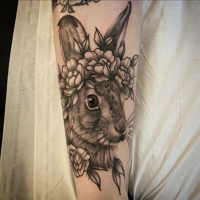 A beautiful black and gray tattoo of a realistic bunny surrounded by intricate floral designs, combining neo-traditional and illustrative styles. By artist Cara Hayward.